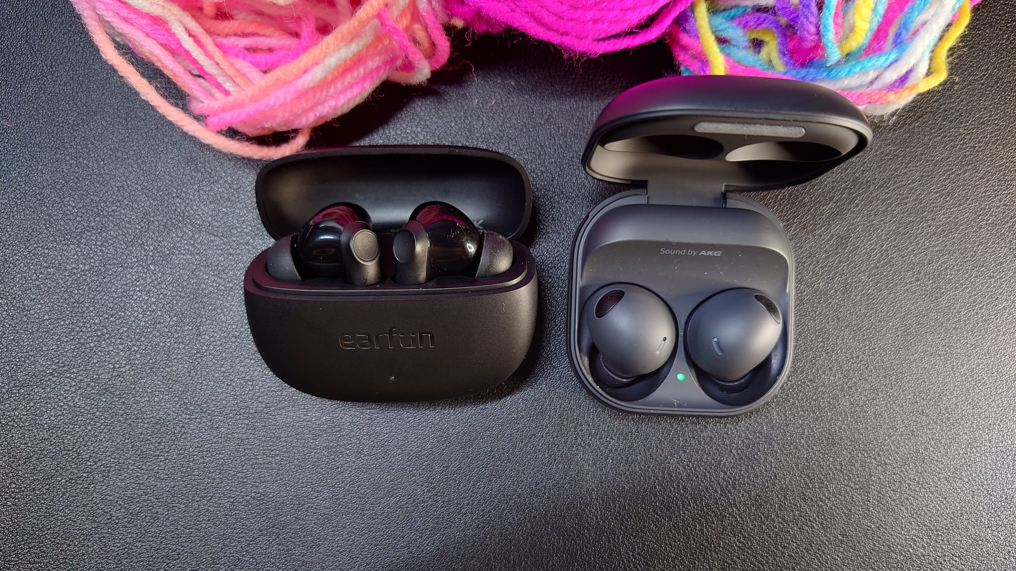 Earfun Air Life and Galaxy Buds 2 Pro, both open in their cases, with 3 shades of pink yarn adorning them at top
