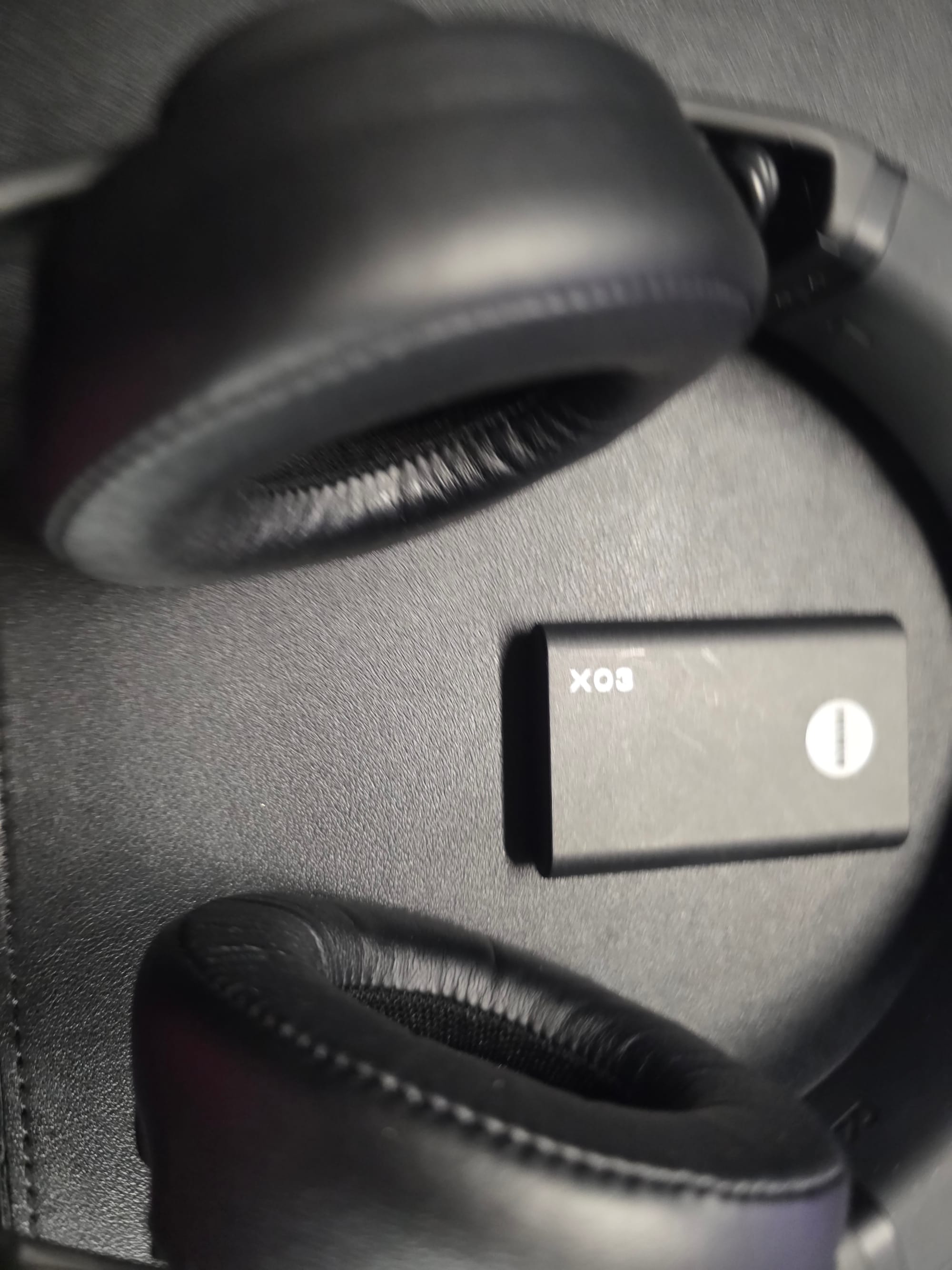 A close look at the AIAIAI TMA-2 studio headphones, which are black with black earcups. The X03 transmitter is also black. Both are lying on a black desk.
